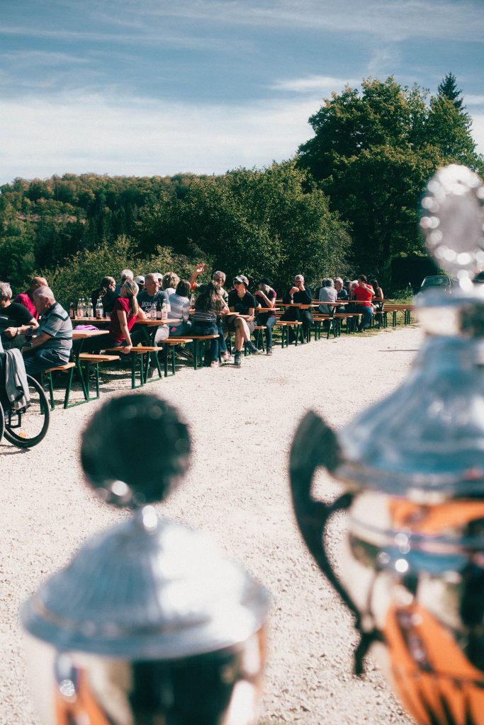 Im Vordergrund: Pokale
Im Hintergrund: Personen sitzen zusammen an Biertischen und reden